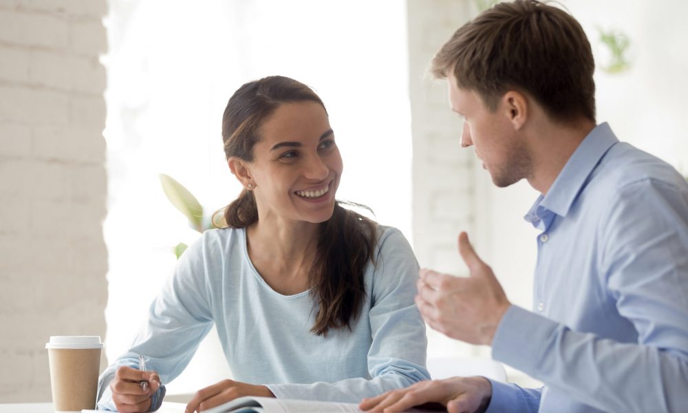Happy mixed race female employee listening to project explanation. Happy mixed race female employee sitting at table, listening to team leader explaining project details, head shot. Young smiling intern involved in educational training of coach mentor at office.